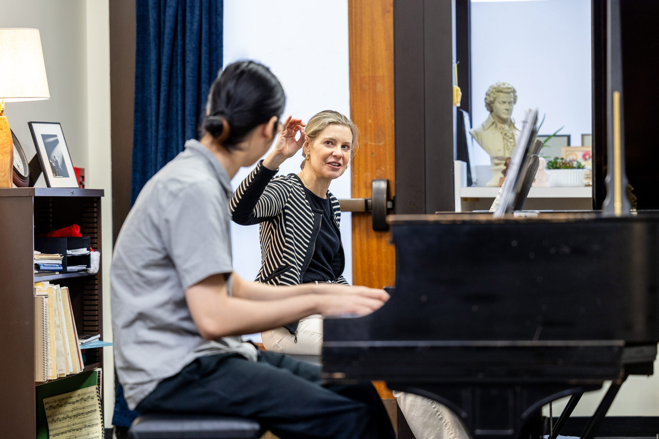 A student sits at a piano in the foreground, blurred while a professor, in focus, is raising their hand in instruction. The room is a filled with a bookcase containing music sheets and photographs framed atop the case and behind the piano we can see a bust of a man in the background