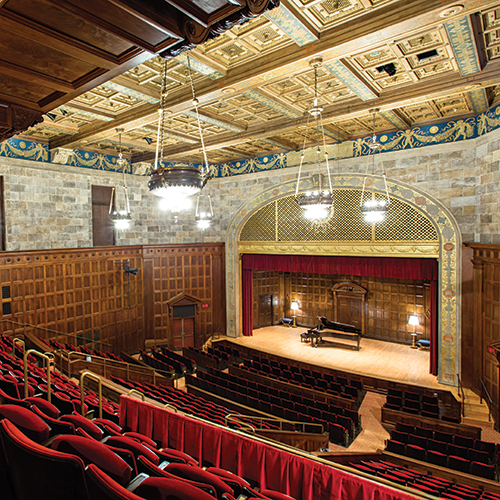A photo looking down at the stage of an empty Kilbourn Hall from upper seats. A piano sits alone on the stage itself.