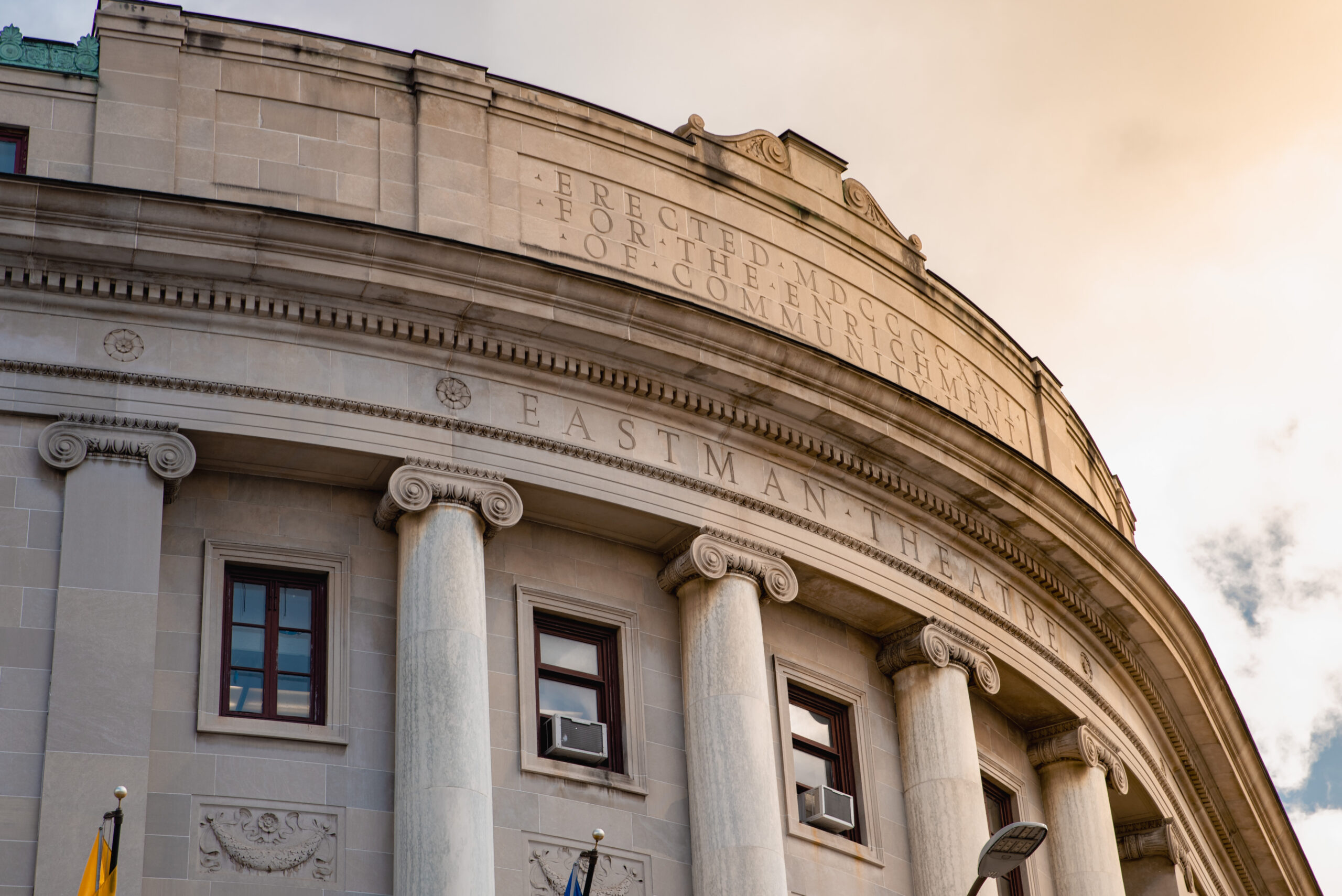 A view of the front of the Eastman Theatre at Eastman School of Music at sunrise. An inscription is visible reading "Erected MDCCCCXXII For the Enrichment of Community life"