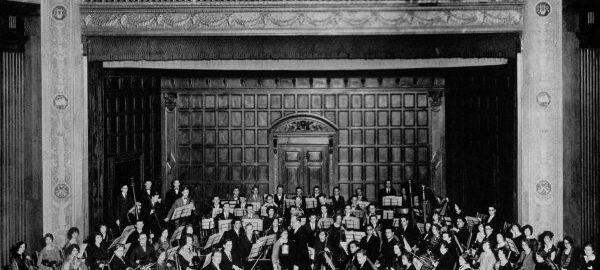 The Eastman School Orchestra (1928). Photograph from The Score (1928). A 70-member orchestra poses with instruments on stage in Kilbourn Hall.