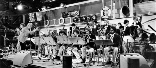 Ray Wright conducts the Eastman Jazz Ensemble during a performance, with scenery depicting a cruise ship in the background.