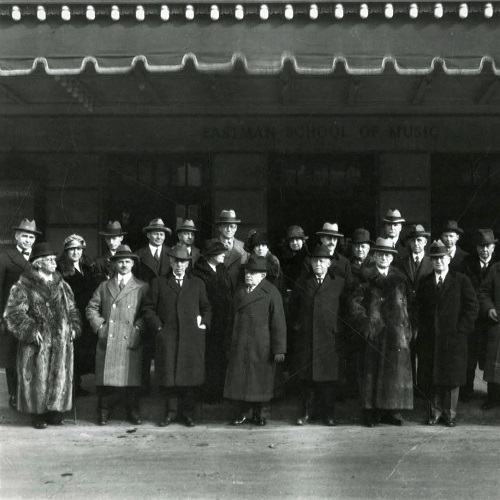 Black and white photo of 22 people, wearing winter coats, standing under the awning outside of the Eastman School of Music's main entrance. Black and white photo of 22 people, wearing winter coats, standing under the awning outside of the Eastman School of Music's main entrance.