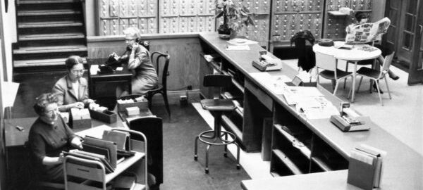 Candid, birds-eye view of the front desk of the old Sibley Music Library, showing Ruth Watanabe and two librarians at work.