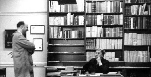 A man in a trench coat stands near Barbara Duncan, seated in her office in front of three full bookshelves. A man in a trench coat stands near Barbara Duncan, seated in her office in front of three full bookshelves.
