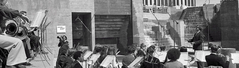 Robert Freeman conducts wind ensemble of Eastman students on a stage in a sunken, concrete plaza.