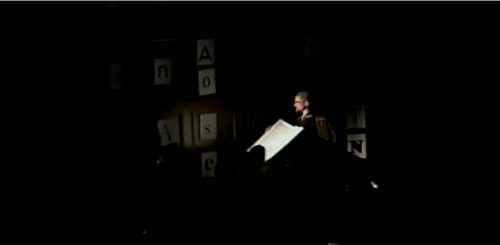 Part II at 32-09 performer holding open a codex to be read from