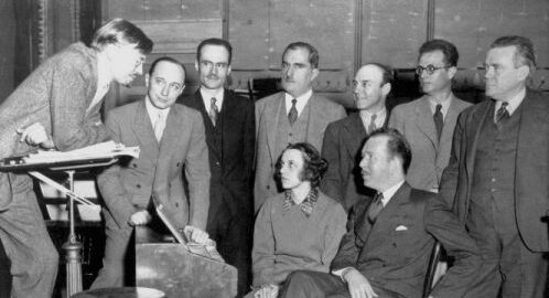 Howard Hanson leans on a podium while speaking to a closely-gathered group of 8 composers in Kilbourn Hall.