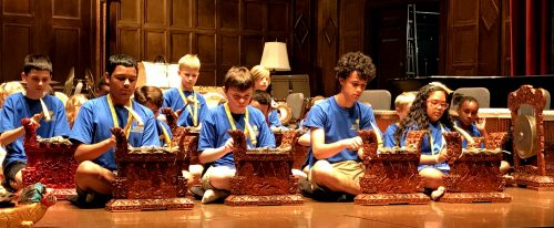 Adventure Music Camp’s gamelan performance Students in blue t-shirts cross-legged on floor playing gamelan instruments.