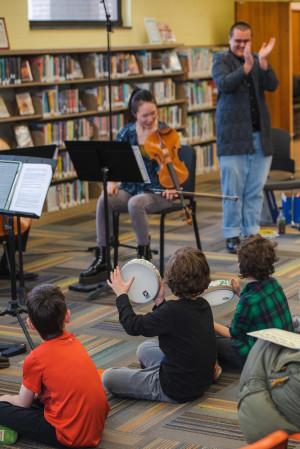 Three children sit on the carpet watching a violinist play. One child clapping a tambourine.