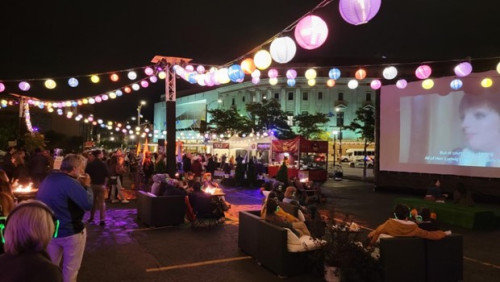 People watch an outdoor movie projected on a screen with a string of lanterns above.