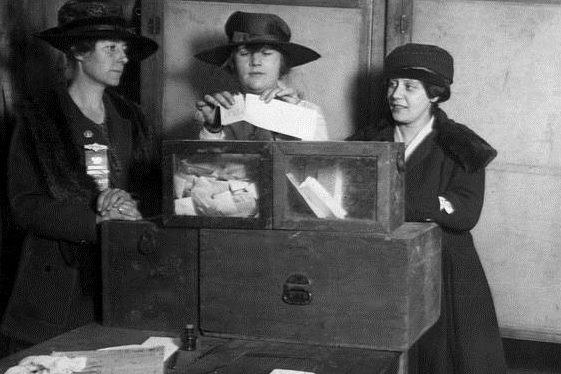 A vintage photo of three Suffragettes around a voting box.