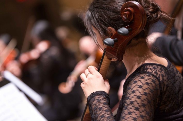 Musicians Unite to Benefit Rochester’s Foodlink A woman in a black dress playing cello in an orchestra setting.